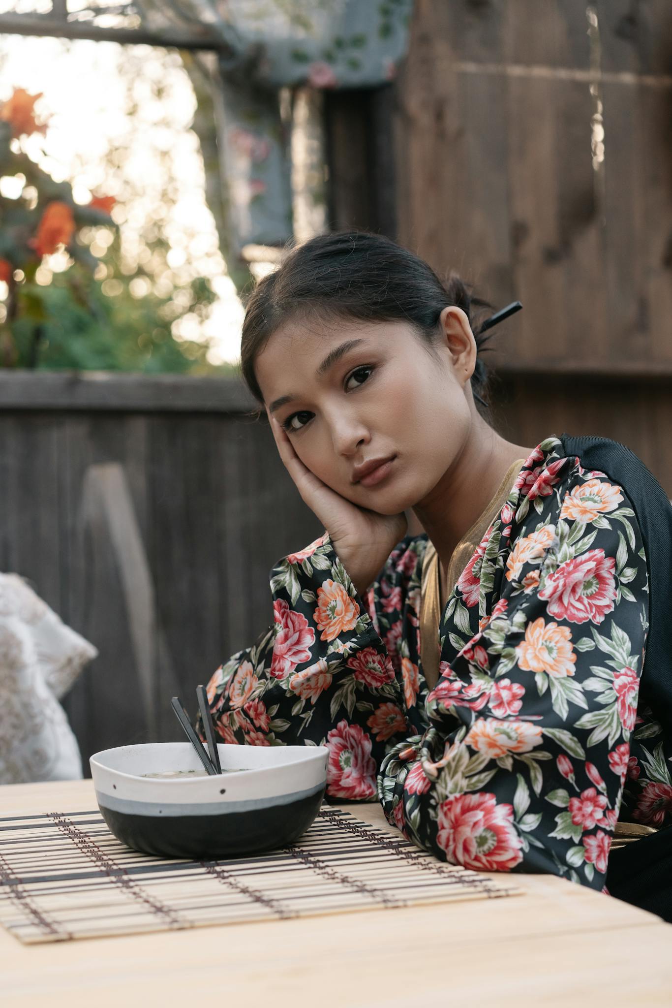 A young woman in a floral kimono sits thoughtfully at a table outdoors.