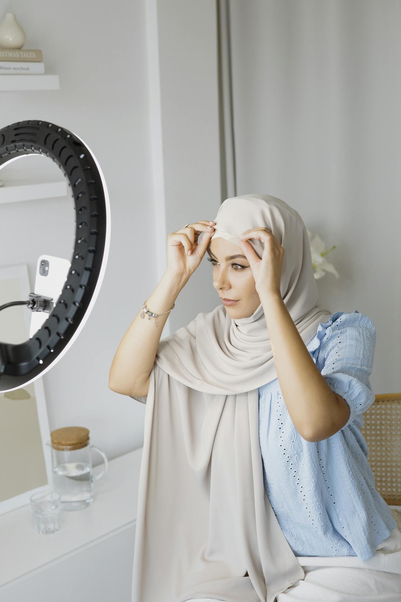 Young woman wearing hijab adjusts smartphone with ring light for social media content creation indoors.